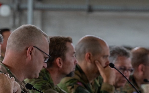 A multinational military briefing or conference is taking place, with numerous uniformed personnel seated along a long table in what appears to be a hangar or large military facility. Personnel are wearing various national military uniforms, including at least one Polish flag patch visible on the leftmost individual. The attendees appear to be attentively listening to a presentation or briefing.