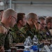 A multinational military briefing or conference is taking place, with numerous uniformed personnel seated along a long table in what appears to be a hangar or large military facility. Personnel are wearing various national military uniforms, including at least one Polish flag patch visible on the leftmost individual. The attendees appear to be attentively listening to a presentation or briefing.