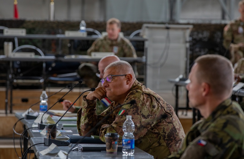 A multinational military briefing or conference is taking place in what appears to be a field operations tent or temporary command facility. Several military personnel from different nations are seated at tables with microphones, water bottles, and documents. The central figure wears an Italian Army patch on his uniform, and another visible soldier appears to wear a Czech Republic flag patch.