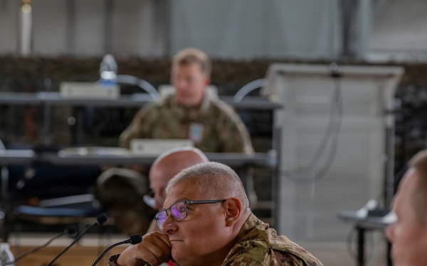 A multinational military briefing or conference is taking place in what appears to be a field operations tent or temporary command facility. Several military personnel from different nations are seated at tables with microphones, water bottles, and documents. The central figure wears an Italian Army patch on his uniform, and another visible soldier appears to wear a Czech Republic flag patch.