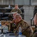 A multinational military briefing or conference is taking place in what appears to be a field operations tent or temporary command facility. Several military personnel from different nations are seated at tables with microphones, water bottles, and documents. The central figure wears an Italian Army patch on his uniform, and another visible soldier appears to wear a Czech Republic flag patch.