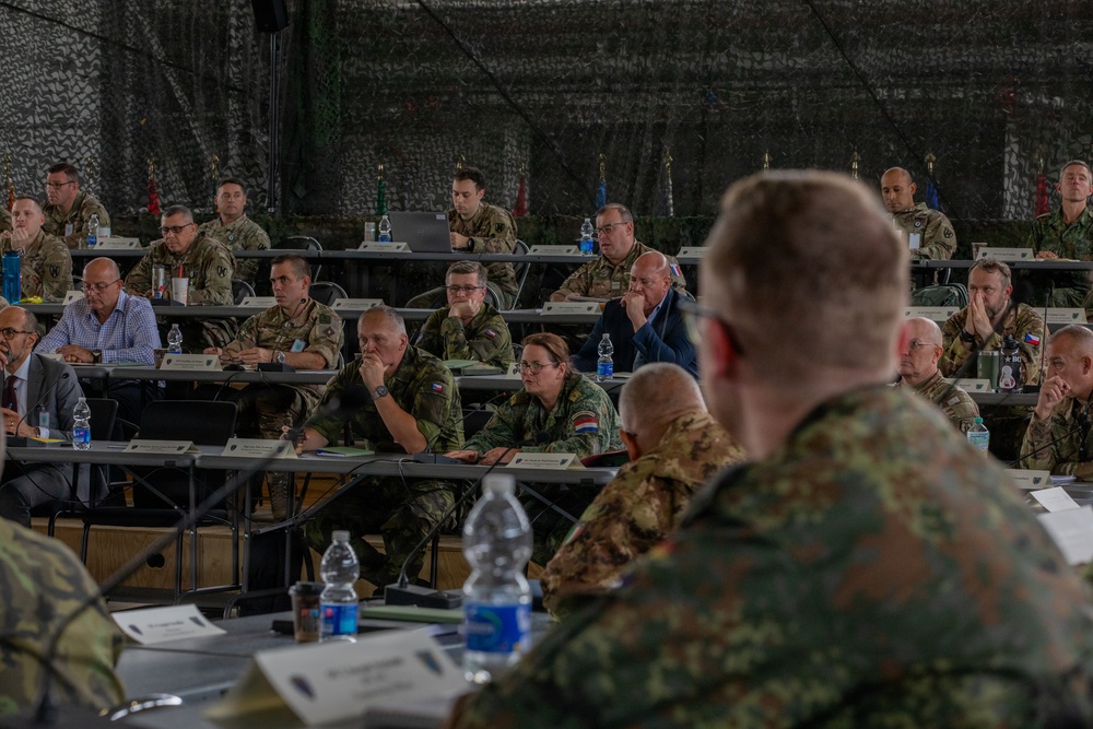 A multinational military briefing or conference is taking place inside a camouflage-netted tent or temporary structure. Personnel in various national military uniforms and some civilian attire are seated at rows of tables, attentively listening to a presenter whose back is visible in the foreground. National flags are visible in the background, indicating a NATO or coalition allied forces gathering.