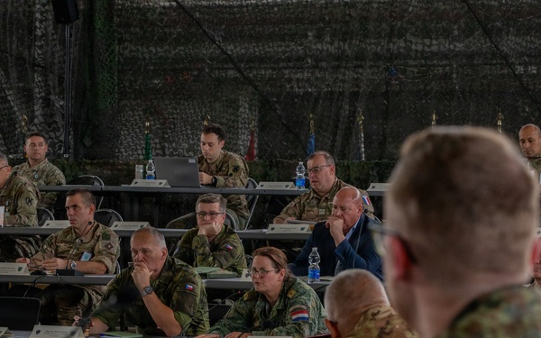 A multinational military briefing or conference is taking place inside a camouflage-netted tent or temporary structure. Personnel in various national military uniforms and some civilian attire are seated at rows of tables, attentively listening to a presenter whose back is visible in the foreground. National flags are visible in the background, indicating a NATO or coalition allied forces gathering.