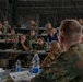 A multinational military briefing or conference is taking place inside a camouflage-netted tent or temporary structure. Personnel in various national military uniforms and some civilian attire are seated at rows of tables, attentively listening to a presenter whose back is visible in the foreground. National flags are visible in the background, indicating a NATO or coalition allied forces gathering.