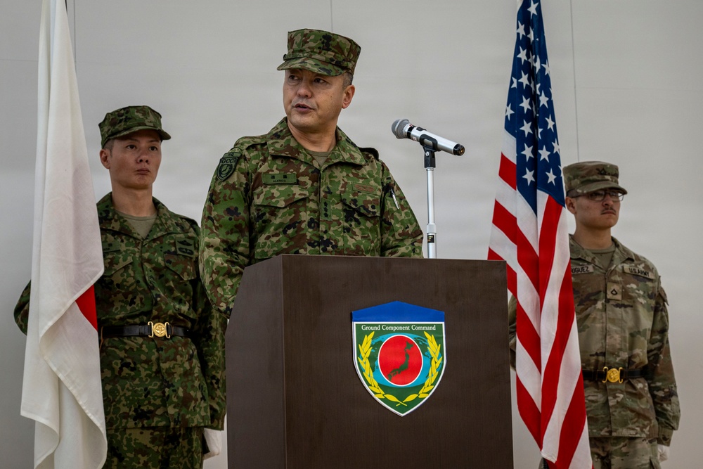 A military officer in Japanese Ground Self-Defense Force (JGSDF) camouflage uniform is speaking at a podium bearing the 'Ground Component Command' insignia. Two other military personnel stand behind him — one in JGSDF uniform and one in U.S. Army OCP (Operational Camouflage Pattern) uniform. The Japanese national flag and the U.S. flag are visible in the background, indicating a joint military ceremony or briefing.