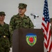 A military officer in Japanese Ground Self-Defense Force (JGSDF) camouflage uniform is speaking at a podium bearing the 'Ground Component Command' insignia. Two other military personnel stand behind him — one in JGSDF uniform and one in U.S. Army OCP (Operational Camouflage Pattern) uniform. The Japanese national flag and the U.S. flag are visible in the background, indicating a joint military ceremony or briefing.