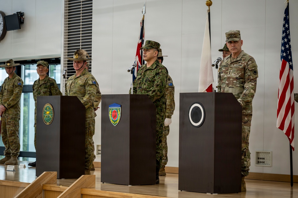A formal multinational military ceremony featuring personnel from at least three nations standing at podiums in an indoor venue. Three podiums display distinct military unit insignia patches, with flags of Australia, Japan, and the United States visible in the background. The event appears to be a joint military command or coordination ceremony.