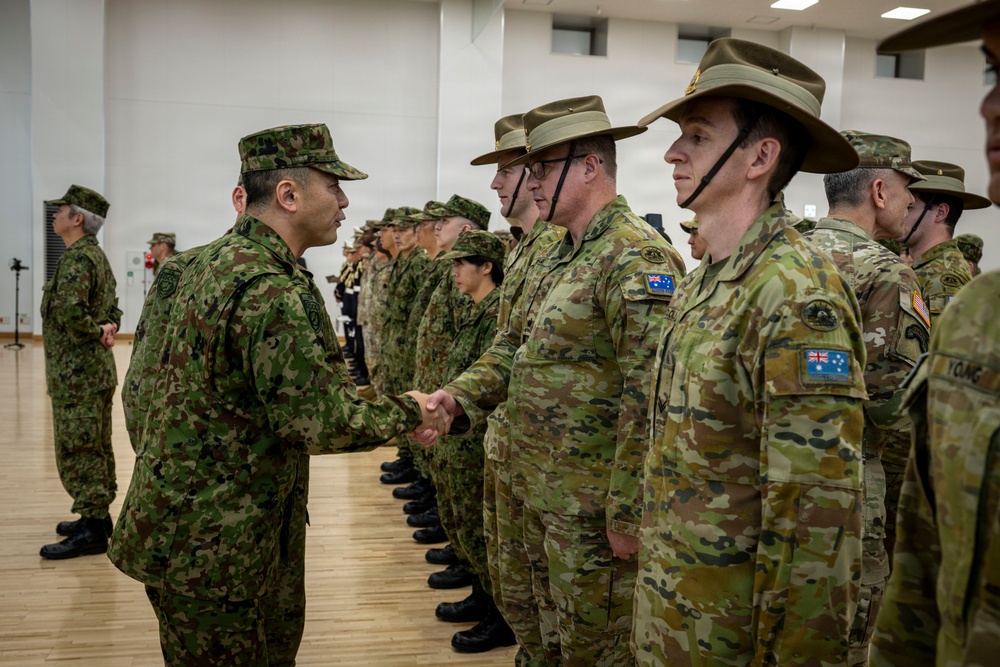 A military ceremony or greeting event is taking place indoors, where a soldier in Japanese Ground Self-Defense Force (JGSDF) camouflage uniform is shaking hands with Australian Army soldiers wearing distinctive wide-brimmed slouch hats and MultiCam uniforms with Australian flag patches. The scene appears to be a formal bilateral military engagement, likely during a joint exercise or official visit. The setting is a large indoor gymnasium or assembly hall with a wooden floor.