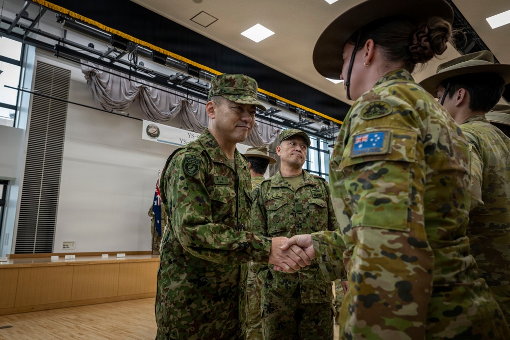 A military ceremony or formal greeting is taking place indoors, likely at a joint military exercise or official event. A senior officer in Japanese Ground Self-Defense Force (JGSDF) camouflage uniform is shaking hands with an Australian Army soldier wearing an Australian flag patch and slouch hat. The setting appears to be a formal hall or auditorium with a banner partially visible in the background.