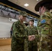 A military ceremony or formal greeting is taking place indoors, likely at a joint military exercise or official event. A senior officer in Japanese Ground Self-Defense Force (JGSDF) camouflage uniform is shaking hands with an Australian Army soldier wearing an Australian flag patch and slouch hat. The setting appears to be a formal hall or auditorium with a banner partially visible in the background.