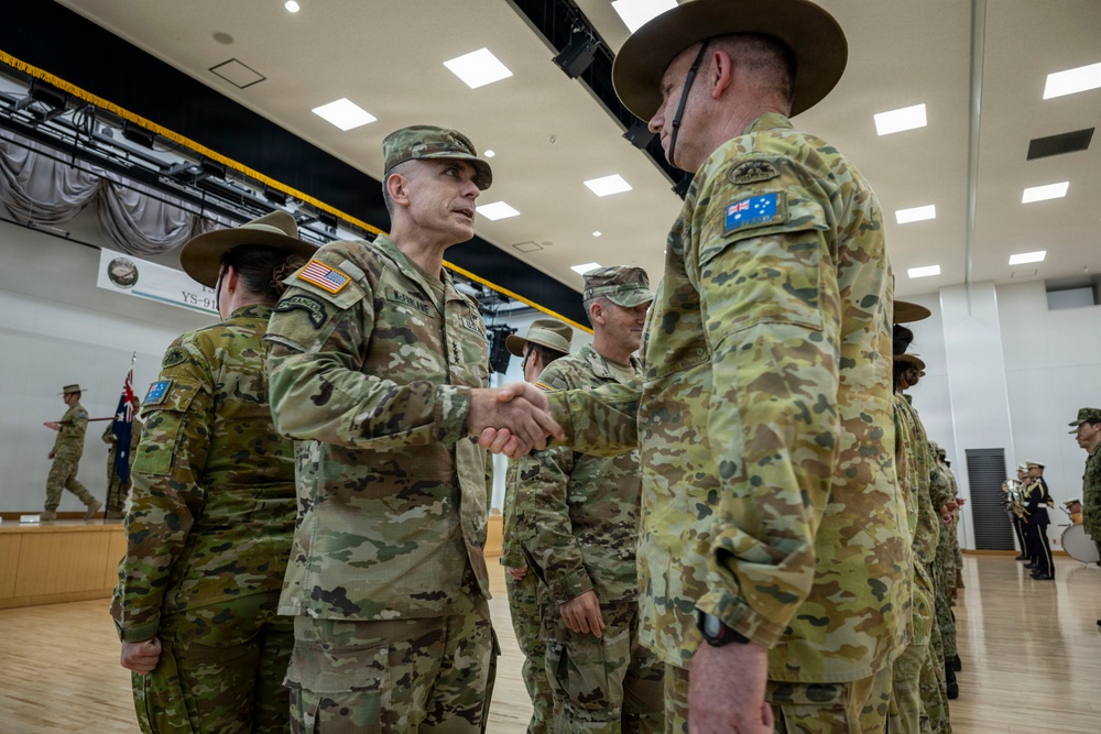 A formal military ceremony showing personnel from the United States Army and the Australian Army exchanging handshakes in what appears to be an indoor venue. The scene depicts an official bilateral military engagement, likely a joint exercise or ceremonial event, with both nations' flags and insignia visible. Multiple uniformed personnel are lined up in formation in the background.