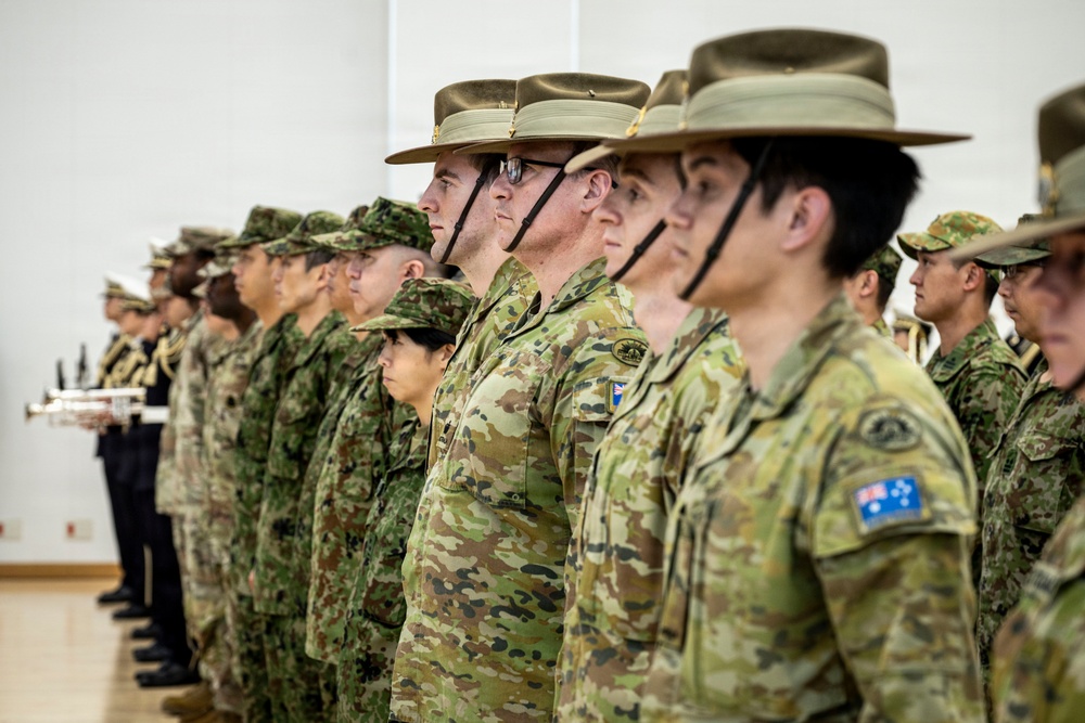 A formal military parade or ceremony featuring personnel from at least two distinct armed forces standing at attention in an indoor venue. Australian Defence Force soldiers are prominently visible wearing distinctive wide-brimmed slouch hats and MultiCam camouflage uniforms with Australian flag patches. Personnel in a different camouflage pattern, consistent with Japan Ground Self-Defense Force uniforms, are also present alongside what appears to be a naval band member in the background.