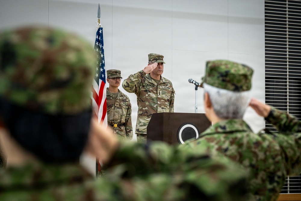 A formal military ceremony is taking place, featuring U.S. Army personnel at a podium saluting alongside an American flag. In the foreground, soldiers wearing a different camouflage pattern — consistent with Japan Ground Self-Defense Force uniforms — are also saluting, suggesting a joint U.S.-Japan military event. The setting appears to be an indoor ceremonial hall.