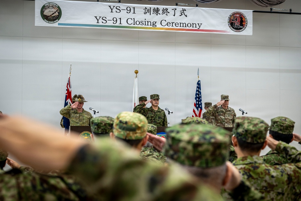 This image depicts the YS-91 Closing Ceremony, a multinational military exercise closing event. Personnel from at least three nations — Australia, Japan, and the United States — are visible on stage saluting, with their respective national flags displayed behind them. The audience, composed primarily of Japanese Ground Self-Defense Force soldiers, is also saluting in unison.