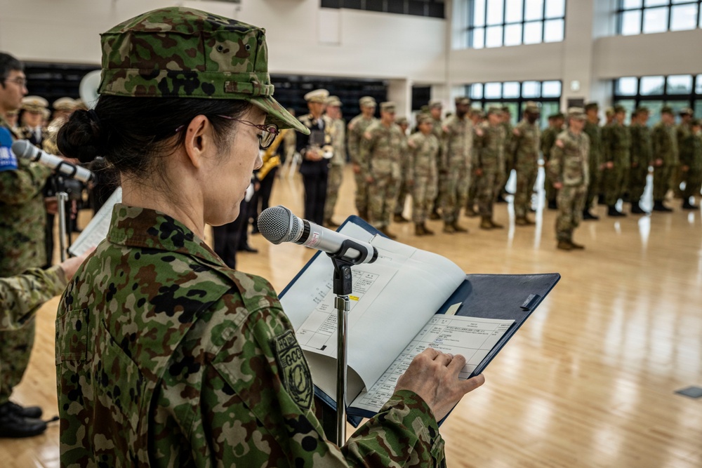 A female military personnel in Japanese Ground Self-Defense Force (JGSDF) camouflage uniform is speaking at a podium microphone while reading from documents on a music stand. Behind her, two formations of soldiers are visible — one in JGSDF uniforms and another in what appears to be U.S. Army MultiCam/OCP uniforms — suggesting a joint military ceremony or exercise. The setting is an indoor gymnasium or drill hall with a hardwood floor.