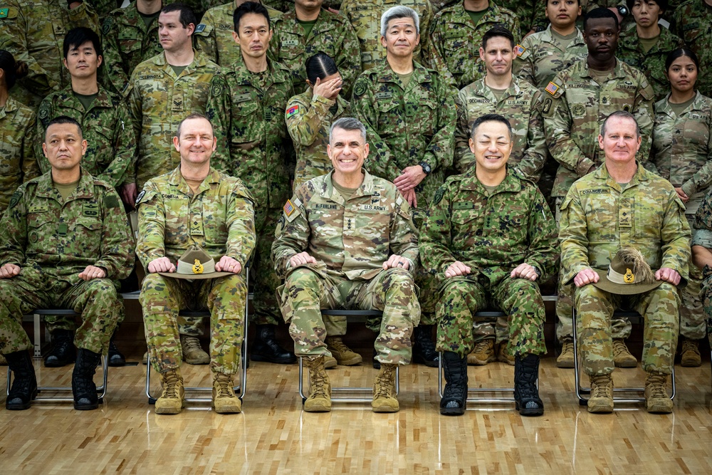 A formal military group photograph featuring personnel from multiple nations, including the U.S. Army, Australian Army, and Japanese Ground Self-Defense Force. Five senior officers are seated in the front row, with a large formation of soldiers standing behind them. The setting appears to be an indoor gymnasium or assembly hall with a hardwood floor.