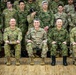 A formal military group photograph featuring personnel from multiple nations, including the U.S. Army, Australian Army, and Japanese Ground Self-Defense Force. Five senior officers are seated in the front row, with a large formation of soldiers standing behind them. The setting appears to be an indoor gymnasium or assembly hall with a hardwood floor.