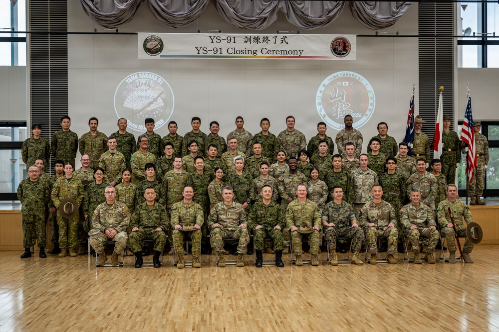 A formal group photograph taken at the YS-91 (Yama Sakura 91) Closing Ceremony, featuring multinational military personnel from the United States, Japan, and Australia. The participants are arranged in four rows in a ceremonial hall, with a banner reading 'YS-91 訓練終了式 / YS-91 Closing Ceremony' displayed prominently behind them. The event appears to be a joint military exercise conclusion ceremony.
