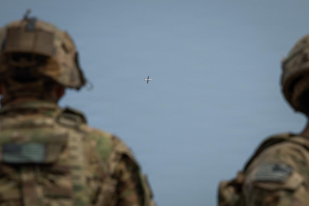 Two military personnel in camouflage uniforms and helmets are observed from behind, watching a small unmanned aerial vehicle (UAV/drone) in flight against a clear blue-gray sky. The soldiers appear to be monitoring or overseeing the drone's operation. The foreground figures are out of focus while the aircraft in the distance is the focal point of their attention.