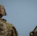Two military personnel in camouflage uniforms and helmets are observed from behind, watching a small unmanned aerial vehicle (UAV/drone) in flight against a clear blue-gray sky. The soldiers appear to be monitoring or overseeing the drone's operation. The foreground figures are out of focus while the aircraft in the distance is the focal point of their attention.