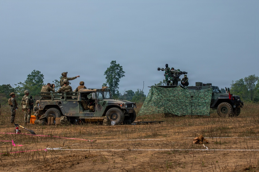 A military field exercise showing two tactical vehicles positioned on an open dirt terrain. Personnel in combat uniforms are gathered around a HMMWV (Humvee) on the left, while a second darker vehicle on the right has a mounted weapon system and camouflage netting deployed. The scene appears to be a joint training exercise with soldiers from at least two different military forces.