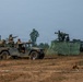 A military field exercise showing two tactical vehicles positioned on an open dirt terrain. Personnel in combat uniforms are gathered around a HMMWV (Humvee) on the left, while a second darker vehicle on the right has a mounted weapon system and camouflage netting deployed. The scene appears to be a joint training exercise with soldiers from at least two different military forces.
