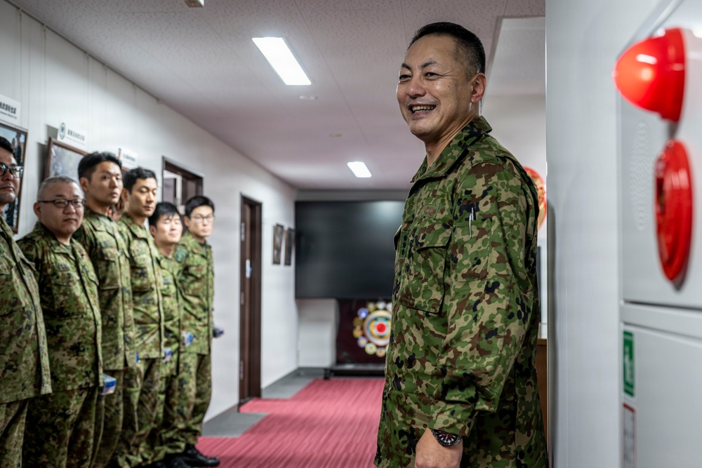 A group of military personnel wearing green camouflage uniforms are gathered in an indoor corridor or hallway. One individual in the foreground is smiling broadly, while others stand in a line along the left wall. The setting appears to be a military facility, with a red fire safety apparatus visible on the right wall.