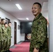 A group of military personnel wearing green camouflage uniforms are gathered in an indoor corridor or hallway. One individual in the foreground is smiling broadly, while others stand in a line along the left wall. The setting appears to be a military facility, with a red fire safety apparatus visible on the right wall.