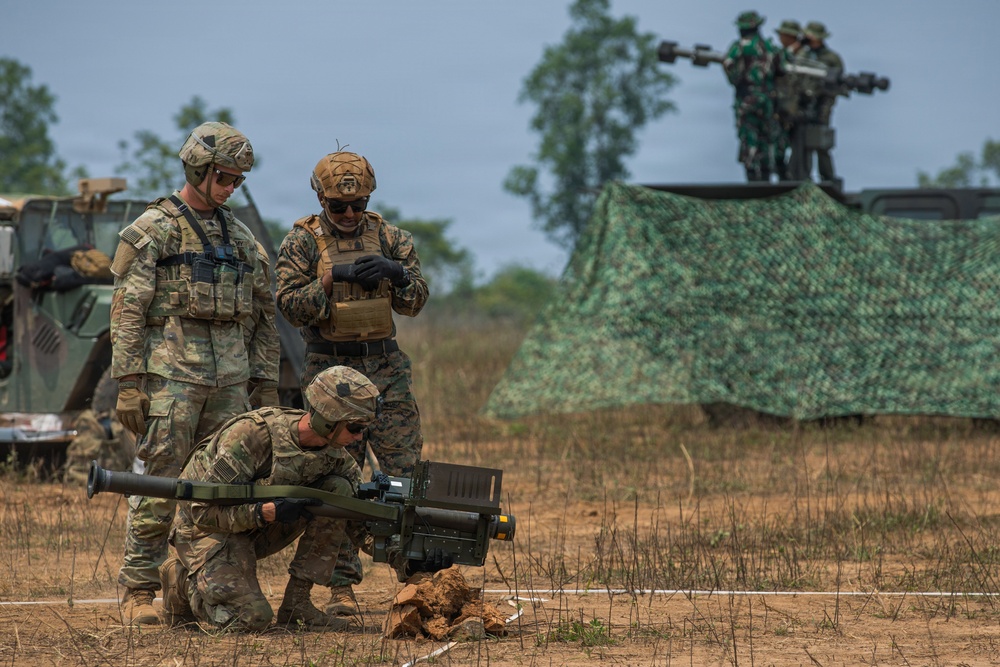 Military personnel in camouflage uniforms are conducting what appears to be a joint training exercise in an open field environment. Soldiers in the foreground are handling a man-portable air-defense or anti-tank weapon system, while additional personnel observe from an elevated position on a vehicle in the background. The presence of different uniform patterns suggests this may be a multinational or inter-service training exercise.