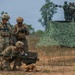 Military personnel in camouflage uniforms are conducting what appears to be a joint training exercise in an open field environment. Soldiers in the foreground are handling a man-portable air-defense or anti-tank weapon system, while additional personnel observe from an elevated position on a vehicle in the background. The presence of different uniform patterns suggests this may be a multinational or inter-service training exercise.