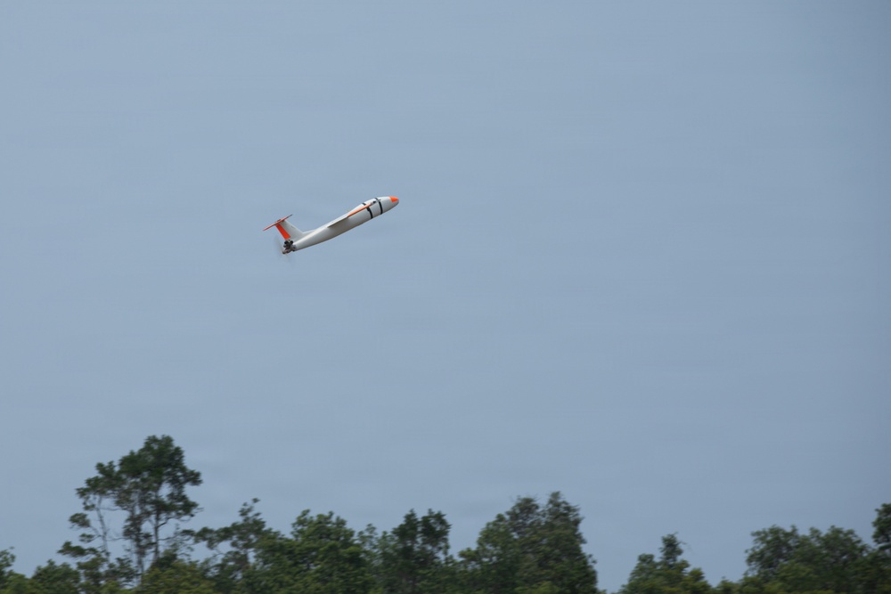 A small white and orange aircraft, appearing to be a radio-controlled (RC) model plane or small UAV, is captured in flight against a clear blue-gray sky. The aircraft is banking at an angle, suggesting it is maneuvering. A treeline of green deciduous trees is visible along the bottom edge of the image.