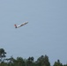 A small white and orange aircraft, appearing to be a radio-controlled (RC) model plane or small UAV, is captured in flight against a clear blue-gray sky. The aircraft is banking at an angle, suggesting it is maneuvering. A treeline of green deciduous trees is visible along the bottom edge of the image.