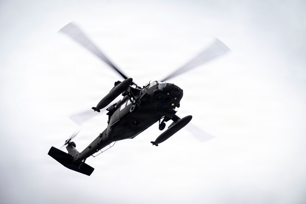 A dark military helicopter is photographed from below against an overcast white sky. The aircraft appears to be in active flight, with main rotor blades visibly blurred from rotation. The low-angle perspective provides a clear view of the helicopter's underside and external components.
