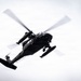 A dark military helicopter is photographed from below against an overcast white sky. The aircraft appears to be in active flight, with main rotor blades visibly blurred from rotation. The low-angle perspective provides a clear view of the helicopter's underside and external components.