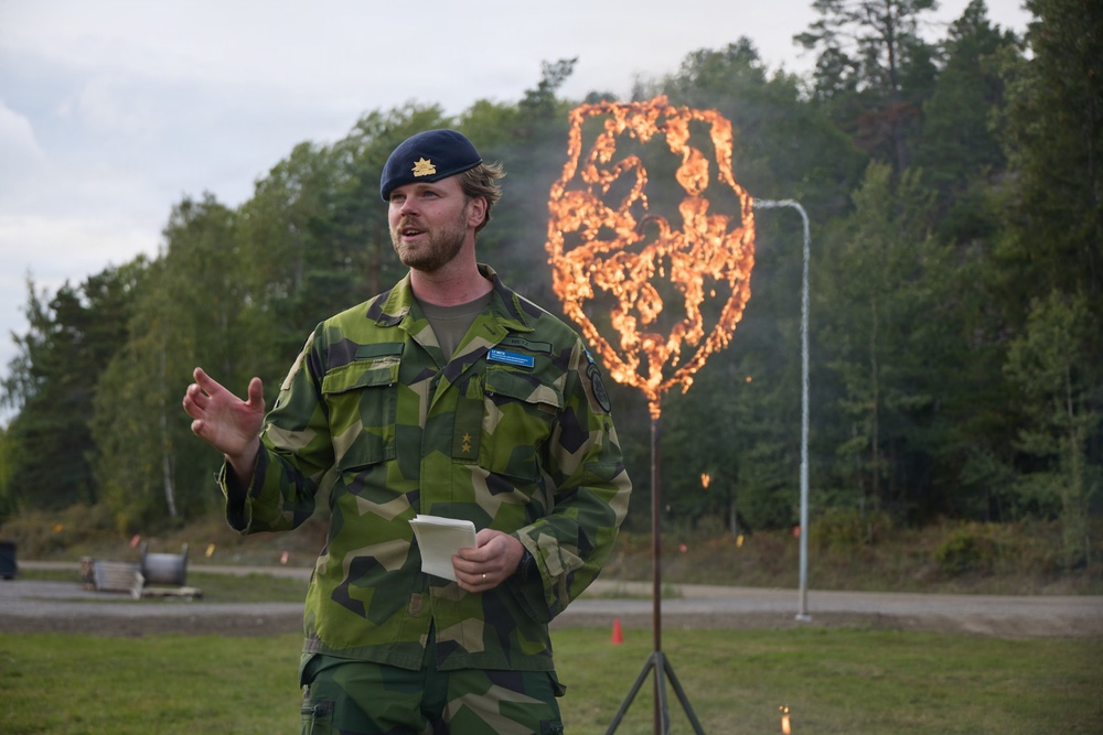 A military officer in camouflage uniform and blue beret is speaking outdoors, holding papers, likely addressing an audience at a ceremony or event. Behind him, a large shield-shaped metal frame is engulfed in flames, mounted on a tripod stand. The setting appears to be a military training ground or ceremonial area surrounded by trees.