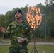 A military officer in camouflage uniform and blue beret is speaking outdoors, holding papers, likely addressing an audience at a ceremony or event. Behind him, a large shield-shaped metal frame is engulfed in flames, mounted on a tripod stand. The setting appears to be a military training ground or ceremonial area surrounded by trees.