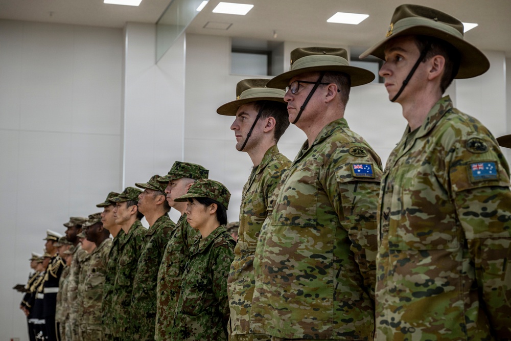 A formal military parade or ceremony featuring personnel from multiple nations standing at attention in a line inside a modern building. Australian Army soldiers wearing distinctive wide-brimmed slouch hats and MultiCam uniforms with Australian flag patches are prominently visible in the foreground, alongside Japanese Ground Self-Defense Force personnel in the middle ground. Additional military personnel in various uniforms, including what appears to be naval dress whites, are visible in the background.