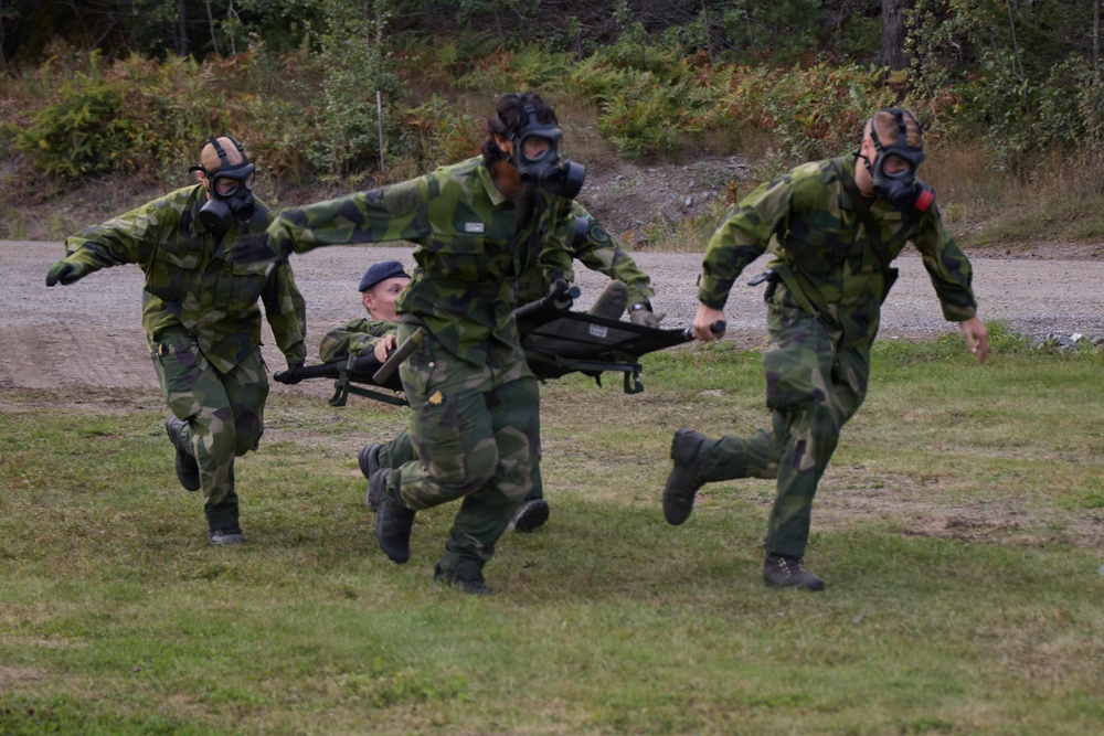 Military personnel in camouflage uniforms and gas masks are conducting a casualty evacuation drill, carrying a soldier on a stretcher at a run. Three soldiers wearing gas masks transport a fourth soldier who is seated/lying on a folding stretcher. The exercise appears to take place on a grassy training area with a gravel road and wooded terrain in the background.