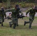 Military personnel in camouflage uniforms and gas masks are conducting a casualty evacuation drill, carrying a soldier on a stretcher at a run. Three soldiers wearing gas masks transport a fourth soldier who is seated/lying on a folding stretcher. The exercise appears to take place on a grassy training area with a gravel road and wooded terrain in the background.