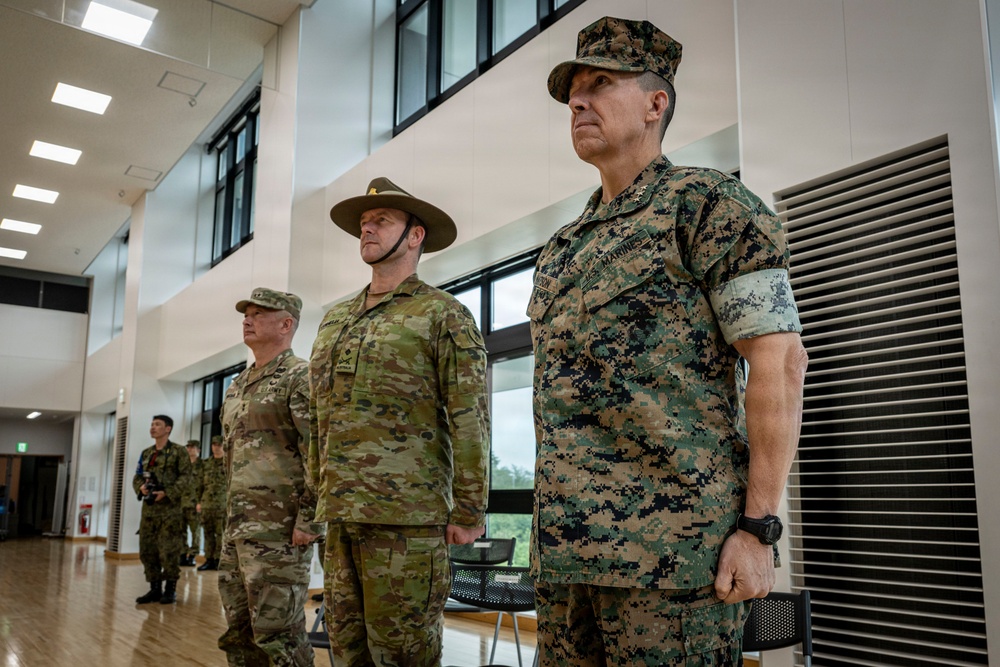 Three military personnel stand at attention in a formal indoor setting, representing at least two different nations' armed forces. The foreground figure wears US Marine Corps MARPAT digital camouflage with visible 'MARINES' tape, while the center figure wears Australian Army multicam uniform with a distinctive wide-brimmed slouch hat. A fourth uniformed individual is partially visible in the background wearing US Army OCP uniform.