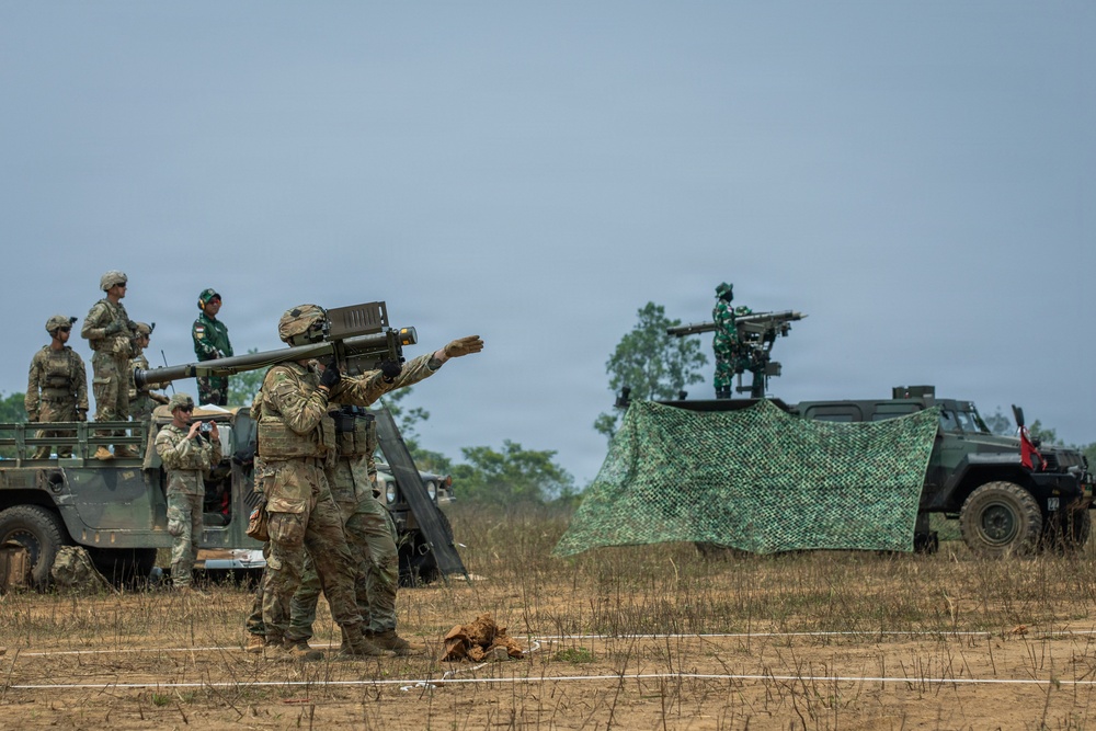 A military exercise or demonstration involving soldiers in multicam/OCP camouflage uniforms and soldiers in green uniforms, likely a joint multinational training event. A soldier in the foreground is shouldering what appears to be a man-portable air-defense system (MANPADS), possibly a Stinger missile launcher. In the background, a military vehicle with a mounted weapon system is positioned under camouflage netting.