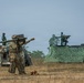 A military exercise or demonstration involving soldiers in multicam/OCP camouflage uniforms and soldiers in green uniforms, likely a joint multinational training event. A soldier in the foreground is shouldering what appears to be a man-portable air-defense system (MANPADS), possibly a Stinger missile launcher. In the background, a military vehicle with a mounted weapon system is positioned under camouflage netting.