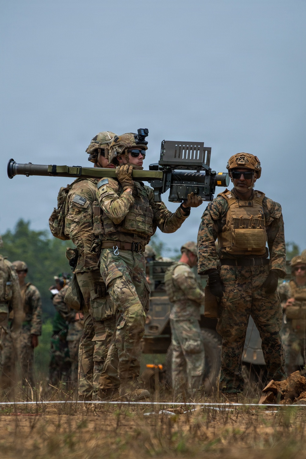 Military personnel in camouflage uniforms are handling what appears to be a man-portable air-defense system (MANPADS) or shoulder-launched missile system during what looks like a training exercise or demonstration. At least two distinct uniform patterns are visible — MultiCam/OCP worn by U.S. Army soldiers and MARPAT worn by a U.S. Marine — suggesting a joint-service or multinational exercise. A military armored vehicle and additional personnel are visible in the background.