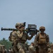 Military personnel in camouflage uniforms are handling what appears to be a man-portable air-defense system (MANPADS) or shoulder-launched missile system during what looks like a training exercise or demonstration. At least two distinct uniform patterns are visible — MultiCam/OCP worn by U.S. Army soldiers and MARPAT worn by a U.S. Marine — suggesting a joint-service or multinational exercise. A military armored vehicle and additional personnel are visible in the background.