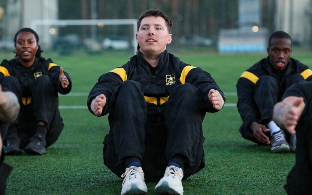 A group of U.S. Army soldiers are performing physical training exercises on an outdoor artificial turf field. They are wearing matching black and gold U.S. Army physical training (PT) uniforms and appear to be doing seated stretching or sit-up exercises. The central soldier is in sharp focus while others are visible on either side.