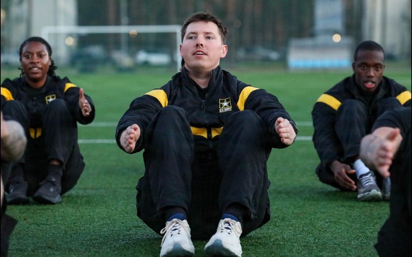 A group of U.S. Army soldiers are performing physical training exercises on an outdoor artificial turf field. They are wearing matching black and gold U.S. Army physical training (PT) uniforms and appear to be doing seated stretching or sit-up exercises. The central soldier is in sharp focus while others are visible on either side.