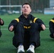 A group of U.S. Army soldiers are performing physical training exercises on an outdoor artificial turf field. They are wearing matching black and gold U.S. Army physical training (PT) uniforms and appear to be doing seated stretching or sit-up exercises. The central soldier is in sharp focus while others are visible on either side.