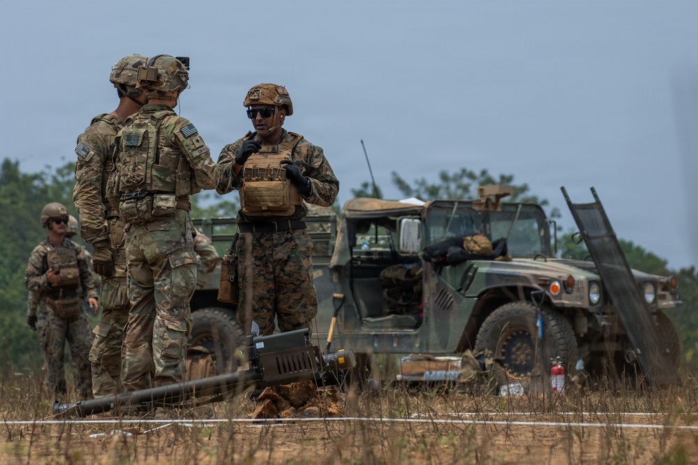 Military personnel in camouflage uniforms are gathered in an open field area near a military Humvee vehicle. Two distinct uniform patterns are visible — MultiCam/OCP (Army) and MARPAT (Marine Corps) — suggesting a joint service operation or exercise. Personnel appear to be coordinating or debriefing near ground equipment on what appears to be a training or operational area.