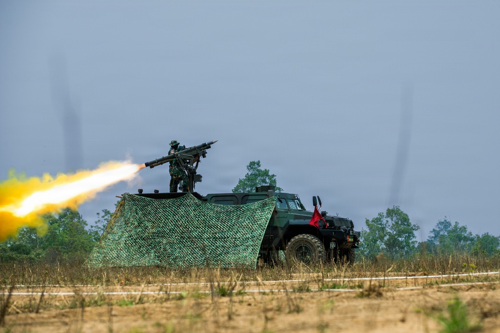 A military vehicle, appearing to be a green armored 4x4 truck, is shown firing a rocket or missile from a launcher mounted on its roof. A soldier is visible operating the weapon system atop the vehicle, which is partially draped with camouflage netting. The scene appears to take place on an open field or training range, with trees visible in the background.
