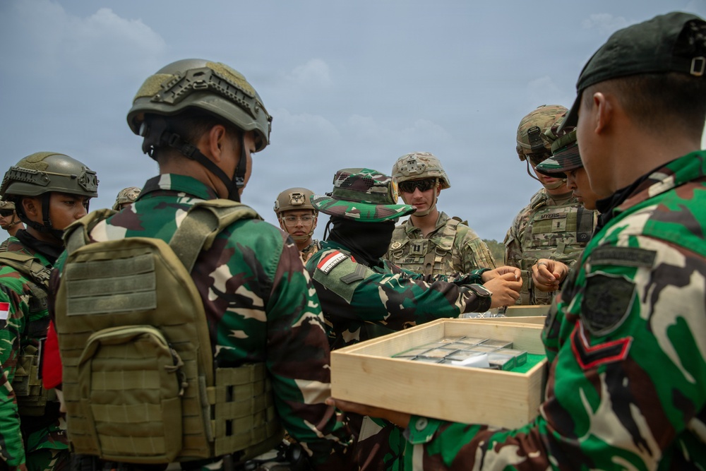 A group of military personnel from two different nations are gathered around a wooden tray containing what appear to be small items, likely ammunition or equipment components. The soldiers include Indonesian Army (TNI) personnel in distinctive green camouflage uniforms with red-and-white Indonesian flag patches, and U.S. Army soldiers in MultiCam/OCP uniforms. This appears to be a joint military training exercise or demonstration.